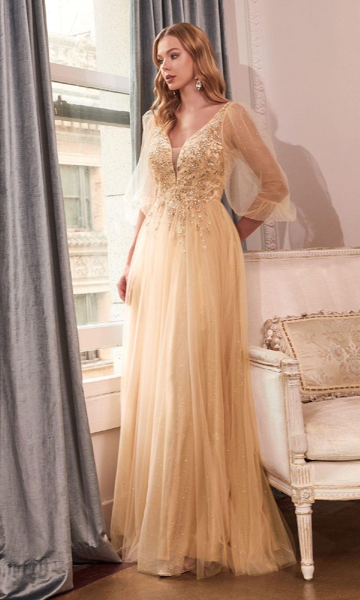 A woman stands indoors in the Ladivine CD0182 sheer-sleeve long prom dress, featuring a beaded v-neck and sequin embellishments, gazing out a window.